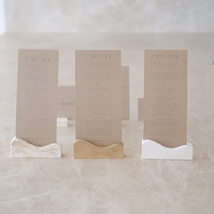 May include: Three rectangular tan paper cards labeled CARE GUIDE, each held upright by a small, wavy-edged base. The bases are finished in a marbled beige, a solid tan, and a solid white, respectively, providing a stable stand for the cards.