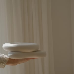 May include: Two stacked, smooth, matte white ceramic ring-shaped objects with rounded edges. The circular forms have a hollow center and a solid, uniform off-white finish, presenting a clean, simple, and sculptural appearance in a neutral tone.