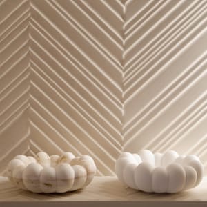 Op de afbeelding: Twee decoratieve kommen met een uniek, afgerond ontwerp. Eén kom is wit met subtiele beige adering, de andere is effen wit. De kommen staan op een lichtgekleurde plank tegen een getextureerde muur met diagonale lijnen.