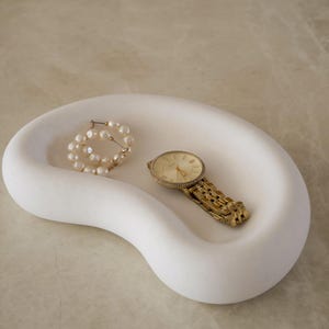 Op de afbeelding: Een witte, niervormige opbergschaal met een gladde, matte afwerking. In de schaal liggen een paar ronde oorringen versierd met kleine witte parels en een goudkleurig analoog horloge met een metalen schakelarmband als horlogeband.