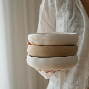 May include: A stack of three round ceramic bowls with irregular, organic edges. The top bowl is off-white with subtle brown marbling, the middle bowl is a solid matte tan, and the bottom bowl is a smooth, solid matte off-white finish.