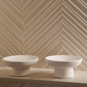 May include: Two white ceramic pedestal bowls with a rounded top and a short, cylindrical base. The bowls are displayed on a light-colored surface against a textured, light-colored wall with a chevron pattern. The bowls are empty.