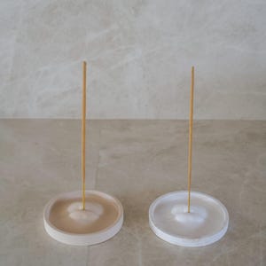 Op de afbeelding: Twee minimalistische wierookhouders. De ene is beige, de andere wit. Elk heeft een cirkelvormige basis met een centrale verdieping om de wierookstok te houden. Een enkele wierookstok staat rechtop in elke houder, klaar voor gebruik.