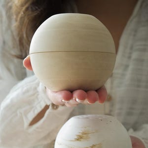 Può includere: Due contenitori decorativi sferici. Il contenitore superiore è di colore beige chiaro uniforme, mentre quello inferiore è bianco con marmorizzazioni marroni. Entrambi sono rotondi con una cucitura al centro.