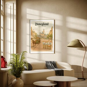 May include: Framed Disneyland Anaheim poster featuring a vintage illustration of the park's entrance, a horse-drawn trolley, and a marching band. The poster is displayed above a cream-colored sofa with a gray throw, and two round coffee tables.