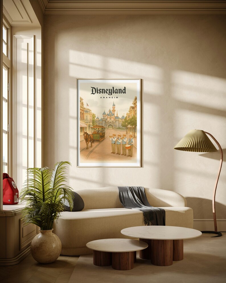 May include: Framed Disneyland Anaheim poster featuring a vintage illustration of the park's entrance, a horse-drawn trolley, and a marching band. The poster is displayed above a cream-colored sofa with a gray throw, and two round coffee tables.