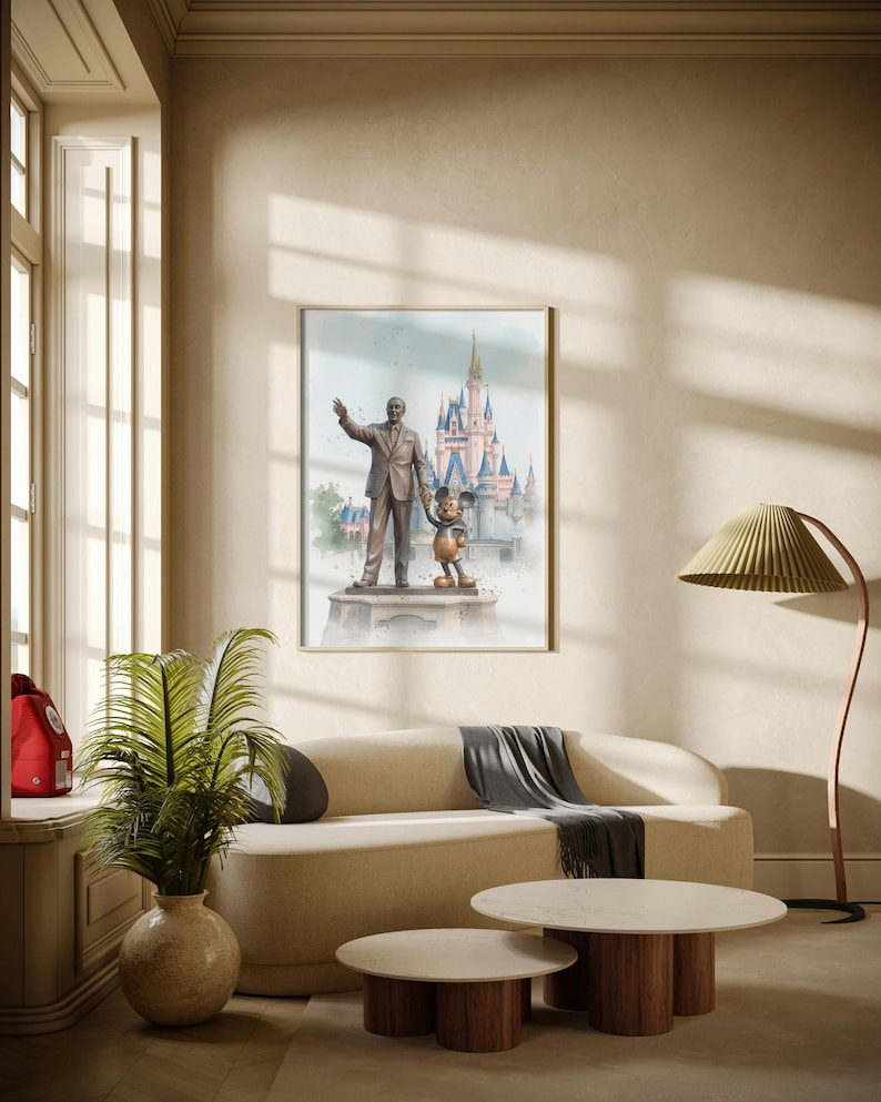 May include: A framed watercolor print featuring a statue of Walt Disney with Mickey Mouse in front of a castle. The artwork is displayed above a beige sofa with a gray throw, and two round coffee tables.