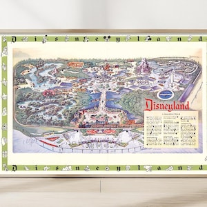 May include: Framed vintage Disneyland map print. The map shows a detailed illustration of the park's layout, with various attractions and landmarks. The print is framed in a light-coloured wooden frame with a green border featuring cartoon characters.