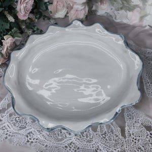 Puede incluir: Una fuente de cerámica blanca con un borde ondulado y festoneado y un ribete azul claro. El plato tiene un acabado brillante y una forma ovalada poco profunda. El fondo incluye flores rosas y encaje.