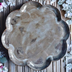 Puede incluir: Una fuente de cerámica hecha a mano con un borde festoneado único. El plato presenta una paleta de colores moteados y terrosos de marrón, gris y blanco, creando una estética rústica pero elegante. Ideal para servir aperitivos o exhibir objetos decorativos.