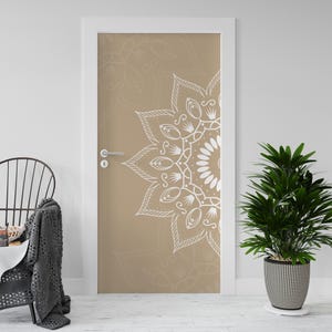 Op de afbeelding: Een deur met een beige achtergrond en een wit mandala-ontwerp. De mandala is een gedetailleerd, symmetrisch patroon. De deur is wit omlijst en heeft een zilveren handgreep. Een plant in pot en een stoel staan op de voorgrond.