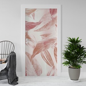 Op de afbeelding: Een deur met een botanische print op grote schaal met oversized bladeren in roze en perziktinten tegen een witte achtergrond. De deur is wit omlijst en heeft een zilveren handgreep. Een plant in pot en een stoel staan op de voorgrond.