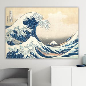 May include: A framed print of "The Great Wave off Kanagawa" by Hokusai. The artwork depicts a large, curling wave in shades of blue and white, with boats and Mount Fuji in the background. The print has a beige background and is displayed on a white wall.