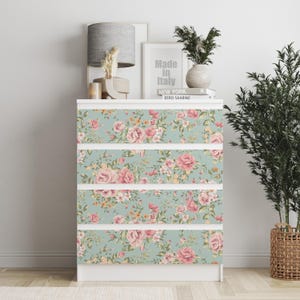 Puede incluir: Una cómoda blanca con cuatro cajones, cada uno adornado con un estampado floral de rosas rosas y hojas verdes sobre un fondo azul claro. La parte superior de la cómoda tiene objetos decorativos, incluyendo una lámpara y una impresión enmarcada que dice "Made in Italy".