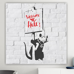 Può includere: Stampa artistica a stencil in bianco e nero su un muro di mattoni bianchi. L'opera presenta un ratto che tiene in mano un cartello con la scritta "WELCOME TO HELL!" in rosso. Il ratto indossa una collana con il simbolo della pace.