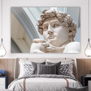 Puede incluir: Una gran impresión en lienzo de la estatua del David de Miguel Ángel, expuesta sobre una cama. La escultura es una vista detallada en primer plano de la cabeza y la parte superior del cuerpo, en tonos blancos y beige. La obra de arte está montada en la pared.