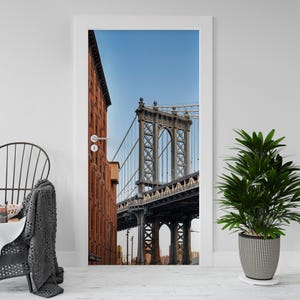 Op de afbeelding: Deur-grote muurschildering met de Manhattan Bridge in New York City. De brug is grijs met gedetailleerd stalen frame, tegen een heldere blauwe lucht. Een rood bakstenen gebouw staat links.