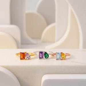 May include: Three gemstone rings in gold, silver, and rose gold. Each ring features two colorful gemstones: orange and purple, purple and green, and blue and yellow. The rings are displayed on a white surface.