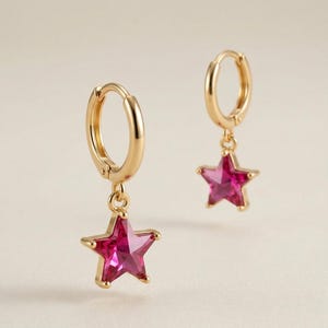 May include: Gold-tone hoop earrings with dangling star-shaped pendants. Each star features a vibrant pink gemstone. The earrings are designed with a simple, elegant style, suitable for everyday wear or special occasions.