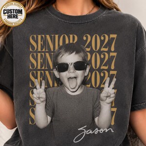 May include: A charcoal gray crew neck t-shirt featuring a black and white photograph of a child wearing sunglasses and making a peace sign with both hands. The text SENIOR 2027 is repeated in gold behind the child, with the name Jason in script below.