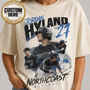 May include: Cream-colored t-shirt featuring a baseball player graphic. The design includes the text "JORDAN HYLAND #27" and "NORTHCOAST BASEBALL" in black and blue. A gold emblem with "CUSTOM DESIGN" and "CUSTOM HERE" is in the upper left.