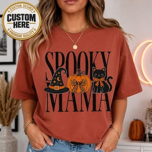 May include: A rust-colored t-shirt featuring a graphic print with the text SPOOKY MAMA. The design includes a black witch hat with yellow stars, an orange pumpkin with a plaid bow, and a black cat wearing an orange plaid bow tie.
