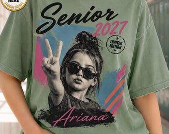 Op de afbeelding: Een saliegroen t-shirt met een grafische print van een persoon met een knotje die een zonnebril draagt en een vredesteken maakt. De tekst luidt Senior 2027, Limited Edition en Ariana. Een ronde badge toont Custom Here Premium Quality.