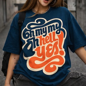 May include: A navy blue t-shirt with the text "Oh my my oh hell YES!" in a retro font. The text is in white, orange, and navy blue. The shirt has short sleeves and a crew neck.