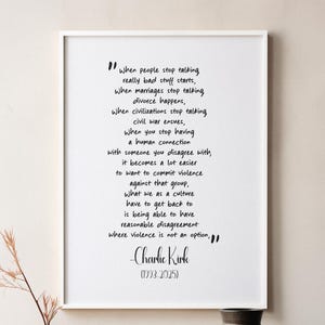 May include: A framed print with a quote by Charlie Kirk, displayed on a wooden table with two ceramic vases and a woven tray. The quote is in black text on a white background. The vases are brown and the tray is a darker brown.