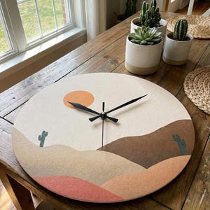 May include: A circular wall clock with a desert landscape design. The clock face has a beige background with an orange sun, brown and tan hills, and green cacti. Black clock hands show the time.