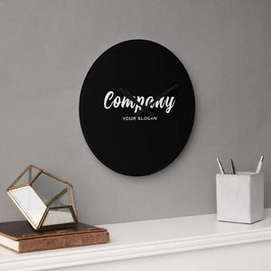 May include: A black, circular wall clock featuring the word "Company" in white script, with "Your Slogan" beneath. The clock hands are black. The clock is mounted on a grey wall. The clock's diameter is approximately 25 cm.