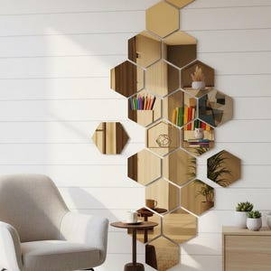 May include: A modern living room with a white wall featuring a honeycomb-shaped mirror arrangement. The mirrors reflect the room's decor, including a gray armchair, a small wooden table, and a wooden cabinet. Decorative items like plants and books are visible.