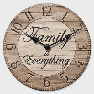 May include: Circular wall clock with a light brown wood grain effect. The clock face displays black numerals and hands, with the words "Family is Everything" in a decorative script. The clock has a dotted border.