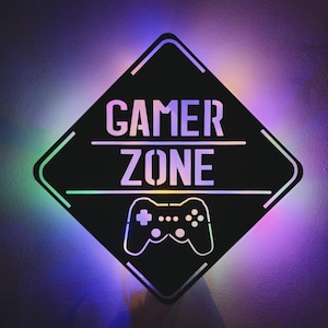 以下が含まれることがあります： ダイヤモンド型で角が丸い黒い金属製の看板。看板には白い文字で「Gamer Zone」と書いてあり、その下に黒いビデオゲームコントローラーが描かれている。看板は虹色の光で照らされている。