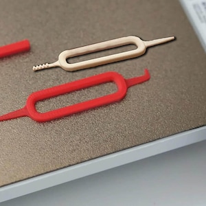 May include: A close-up shot of three small, plastic tools. Two are a red color, and one is off-white. The tools have a rectangular shape with a loop in the middle and pointed ends. One red tool has a hook-shaped end.