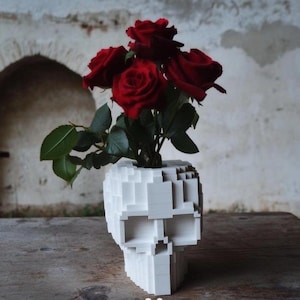 Könnte beinhalten: Eine weiße, blockförmige Schädelvase mit roten Rosen. Die Vase hat ein pixelartiges Design. Die Rosen haben grüne Blätter, und die Anordnung befindet sich auf einem Holztisch.