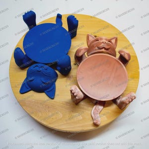 May include: A wooden serving tray with two cat-shaped coasters and one cat-shaped holder. The coasters are blue and the holder is brown. The cat figures have outstretched paws and are lying on their backs. The tray is light brown.