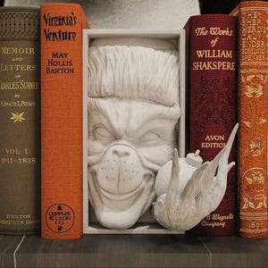 Pode incluir: Uma composição decorativa de estante com livros antigos e uma escultura branca de um rosto de cão. Os livros incluem títulos como "Memoir and Letters" e "Virginia's Venture". Uma escultura de pássaro branco também está presente.