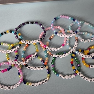 Könnte beinhalten: Eine Sammlung von bunten Perlenarmbändern, die jeweils einen Namen mit weißen Buchstabenperlen buchstabieren. Namen sind u.a. Jasmine, Ursula, Tinkerbell, Moana. Die Armbänder liegen auf einer hellblauen Oberfläche.