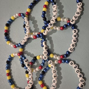 May include: A collection of beaded bracelets. Each bracelet features white letter beads spelling out words such as "Fantasy" and "Adventure". The bracelets are made with blue, yellow, red, white, and black beads. They are displayed on a light blue surface.