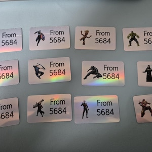 Può includere: Adesivi rettangolari con illustrazioni di supereroi, tra cui Thor e Spider-Man. Ogni adesivo ha il testo "From" e un numero. Gli adesivi hanno un effetto olografico.