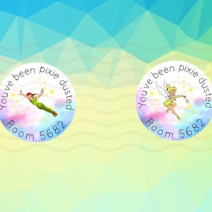 Puede incluir: Dos pegatinas circulares con temática de dibujos animados. Una presenta a Peter Pan, la otra a Campanilla. Ambas tienen el texto "You've been pixie dusted" y "Room 5682" en un diseño circular. El fondo es un degradado de azul, verde y amarillo.