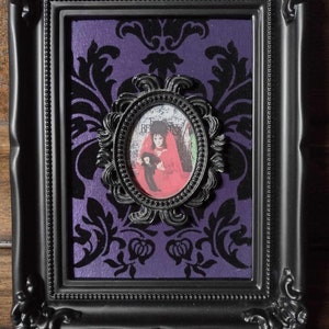 Puede incluir: Una imagen enmarcada en negro con un fondo morado y una imagen ovalada en blanco y negro de una mujer con un vestido rojo sosteniendo una muñeca. La imagen ovalada tiene un marco negro adornado.