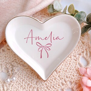 May include: A heart-shaped, white ceramic trinket dish with a gold rim. The dish features the name "Amelia" in pink script, along with a bow design. A pair of pearl earrings rests near the dish, suggesting a jewelry theme.