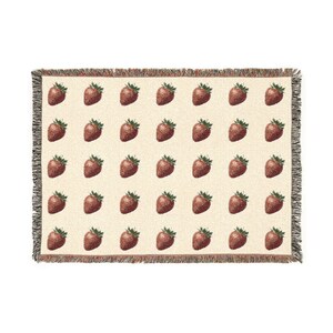 Könnte beinhalten: Eine rechteckige, gewebte Wohndecke mit einem sich wiederholenden Muster aus roten Erdbeeren auf cremefarbenem Hintergrund. Die Decke hat an allen vier Kanten einen dekorativen Fransenrand, der eine mehrfarbige Fadenstruktur aufweist.