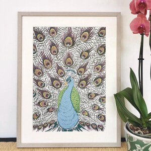 May include: A framed print of a peacock with its feathers displayed. The peacock is depicted in blue and green, with detailed feather patterns in brown and gold. The frame is light grey. A pink orchid is to the right of the framed print.