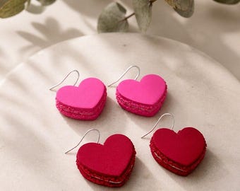 Pode incluir: Dois pares de brincos em forma de coração que lembram macarons. Um par é rosa choque e o outro é de um vermelho carmesim profundo. Cada brinco possui um gancho de metal prateado e uma borda texturizada que imita a camada de um biscoito.