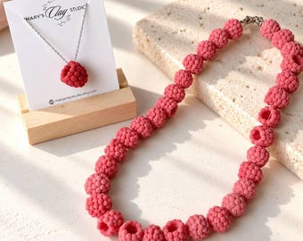 Pode incluir: Um colar com uma série de contas texturizadas em forma de framboesa na cor rosa escuro. Ao lado, um cartão de apresentação branco com o texto 'Mary's Clay Studio' contém um pingente de framboesa a condizer numa corrente prateada fina.