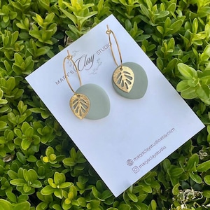 Könnte beinhalten: Ein Paar salbeigrüne, kreisförmige Tonohrringe mit goldfarbenen Metallblattanhängern an goldenen Creolen. Die Ohrringe sind auf einer weißen Karte mit dem Text Mary's Clay Studio, marysclaystudio.etsy.com und marysclaystudio befestigt.