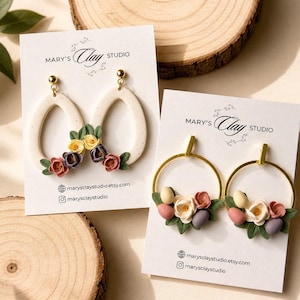 Könnte beinhalten: Zwei Paar Ohrringe auf weißen Präsentationskarten mit der Aufschrift Mary's Clay Studio. Ein Paar zeigt cremefarbene, gesprenkelte Tropfenformen mit kleinen Tonblumen. Das andere Paar besteht aus goldfarbenen Reifen mit Tonblumen und Eiern.